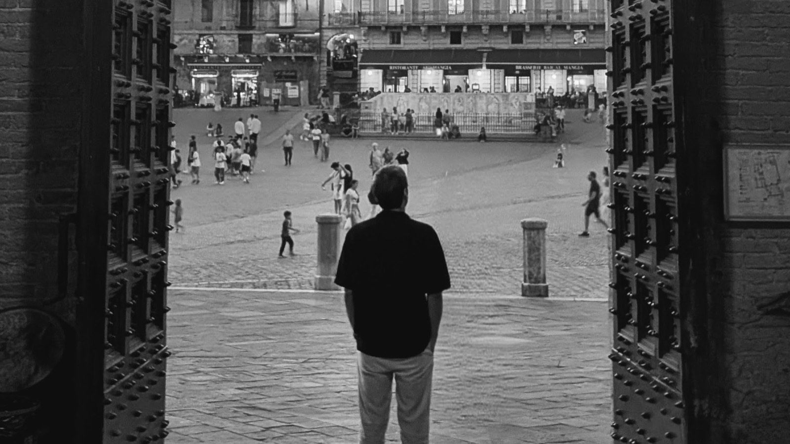 A man stands in a doorway looking out over a busy square, in black and white
