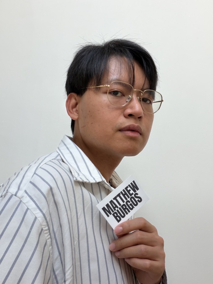 Matthew Burgos holding a card with his name, in a striped shirt against a white wall